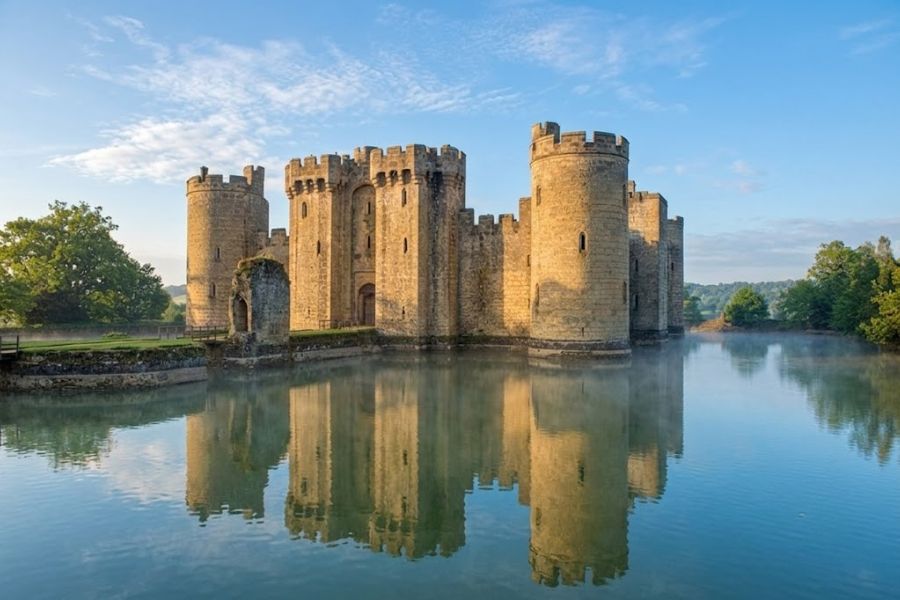 Stone castle with round towers rises from still water under a pale blue sky. Its full reflection in the moat captures the classic look of English castles in the countryside.