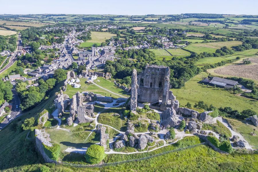 Aerial view of a ruined hilltop castle with broken stone walls and a central tower above a village and patchwork fields. The wide landscape shows how many English castles were built to watch over nearby towns and farmland.