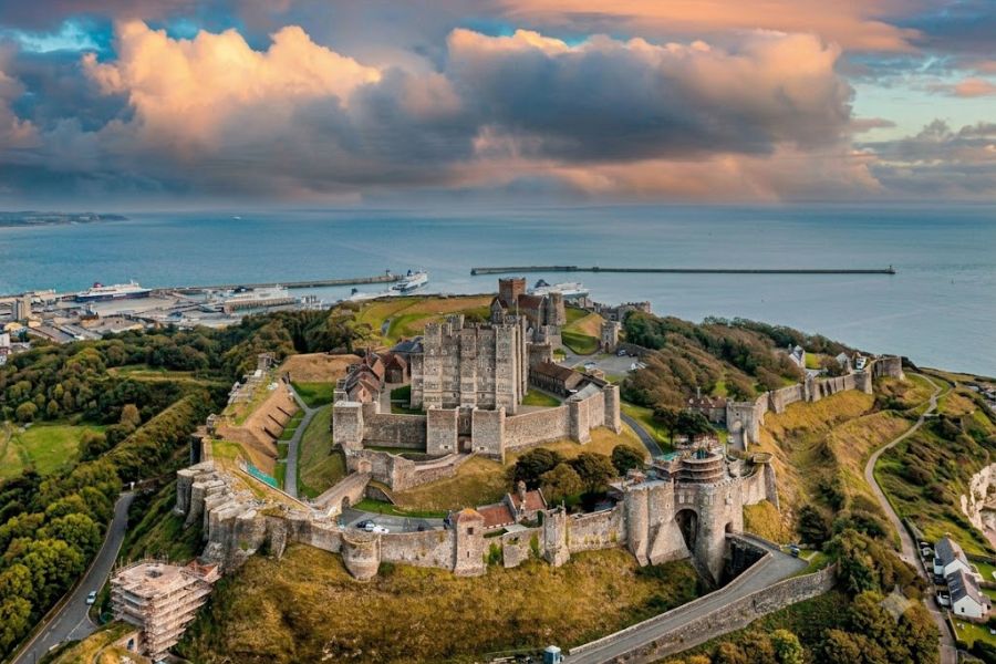 Massive stone fortress sits on a high coastal headland with the sea and harbor stretching out behind it. The layered walls and hilltop setting highlight the defensive scale of English castles.
