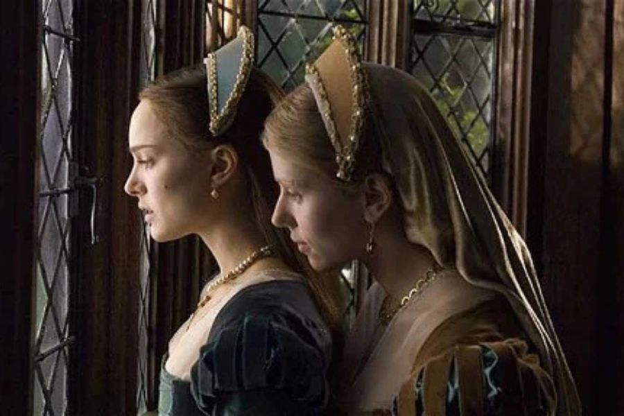 Two women in Tudor gowns and angular French hoods stand closely together beside a leaded glass window. The dramatic scene evokes the courtly setting of stories about Anne Boleyn.
