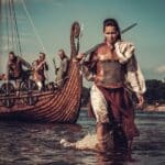 Woman dressed in chainmail and a fur cloak wades ashore carrying a sword while a Viking style longship and armed crew wait behind her in shallow water. This dramatic reenactment illustrates the role of Anglo Saxon women in early medieval life and challenges the idea that women were only passive members of these societies.