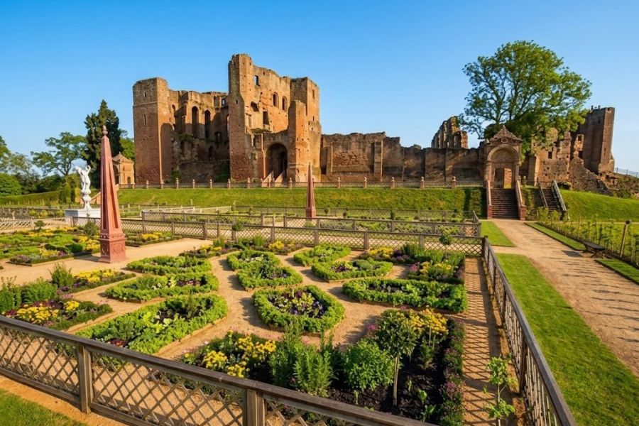 Formal knot garden blooms in the foreground with the ruins of a large red stone castle behind it. The contrast between neat flower beds and weathered walls gives this English castle a romantic feel.