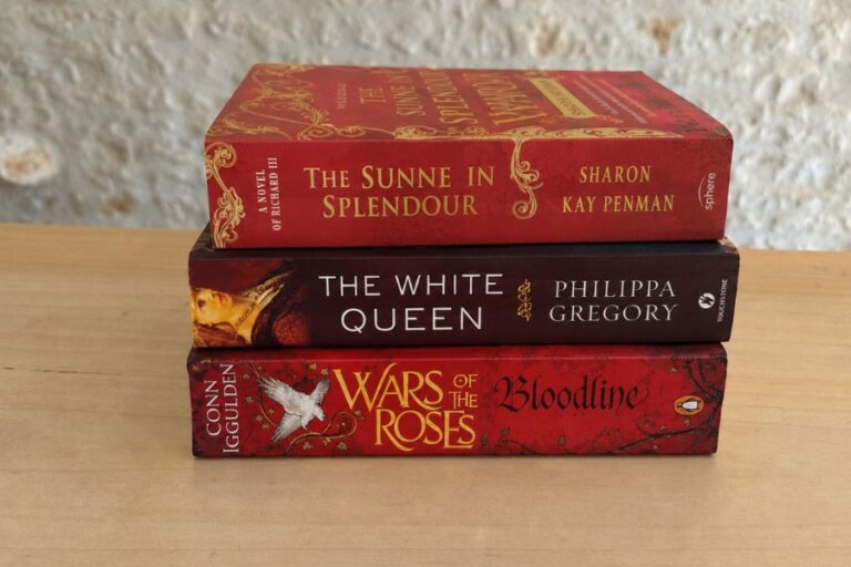 Three historical fiction books about the Wars of the Roses stacked on a wooden table against a pale stone wall. The visible spines read "The Sunne in Splendour. Sharon Kay Penman." "The White Queen. Philippa Gregory." and "Wars of the Roses. Bloodline. Conn Iggulden."