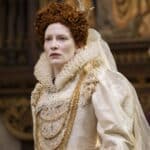 A pale woman with tightly curled red hair wears an elaborate white gown with a high lace ruff and layered pearl necklaces. Her light complexion and carefully styled appearance reflect the beauty ideals of a royal court.