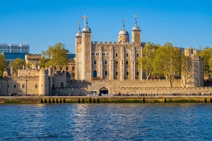 Riverside view of a large stone fortress with square walls and four white topped towers. Trees and the water in front frame one of the most recognizable English castles in London.