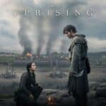 Promotional artwork for The Uprising showing a hooded man holding a curved blade standing over a kneeling woman as a fortified castle burns in the background beneath dark, stormy skies. The dramatic medieval scene reflects the conflict and tension at the heart of The Uprising, with the title "THE UPRISING" displayed across the top.
