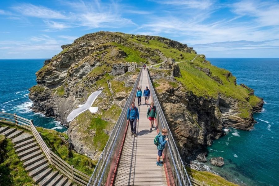 Visitors walk across a narrow footbridge to a rocky island with steep green cliffs and blue sea on both sides. This dramatic coastal path leads to castle ruins and shows a wilder side of English castles.