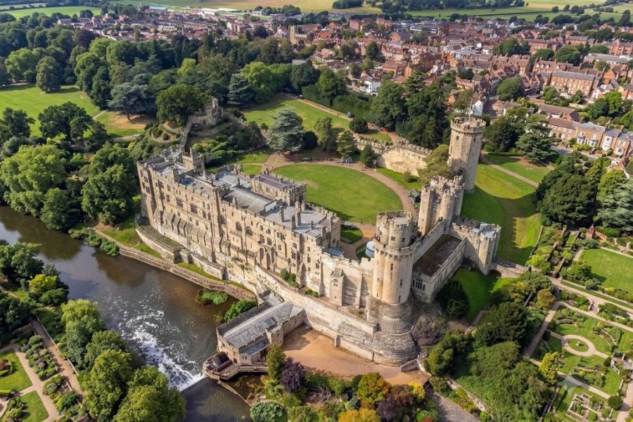 Aerial view of a large stone castle with towers and curtain walls beside a river and a town. Green lawns and formal gardens surround the complex making it one of the most picturesque English castles.