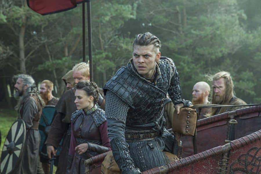 An armored Viking warrior leans forward from a decorated wooden vessel while several fighters gather behind him in a forest clearing. The tense scene evokes stories of Ivar the Boneless preparing for conflict.