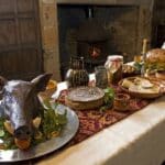Tudor England inspired banquet table with a boar head centerpiece, pie, bread, cheese, preserves, nuts, and a roasted bird arranged near a stone fireplace. The setting suggests a historic feast inside a wood paneled hall.