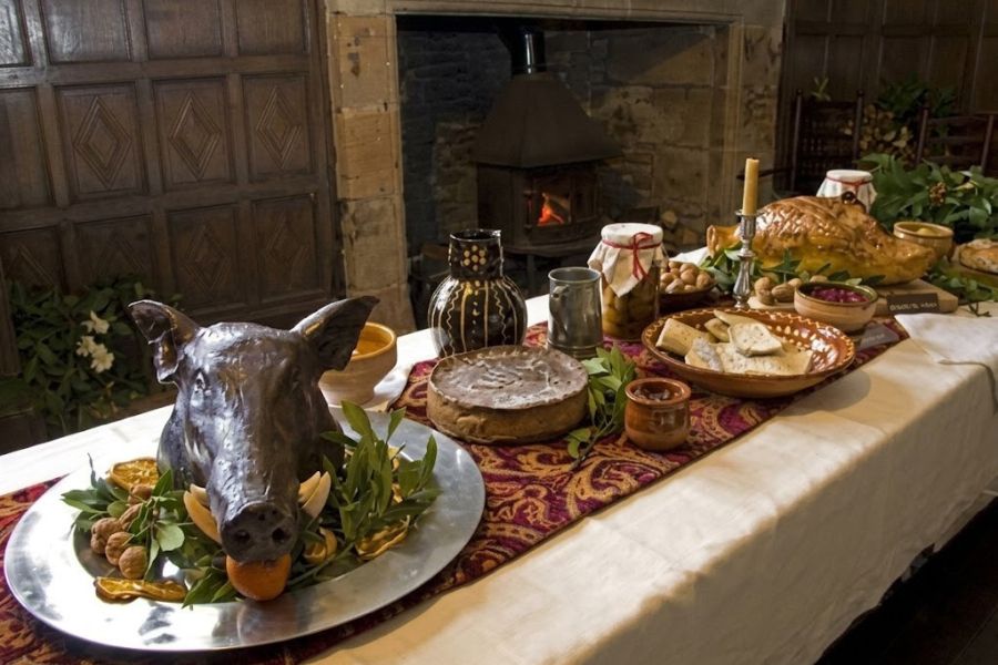 Tudor England inspired banquet table with a boar head centerpiece, pie, bread, cheese, preserves, nuts, and a roasted bird arranged near a stone fireplace. The setting suggests a historic feast inside a wood paneled hall.