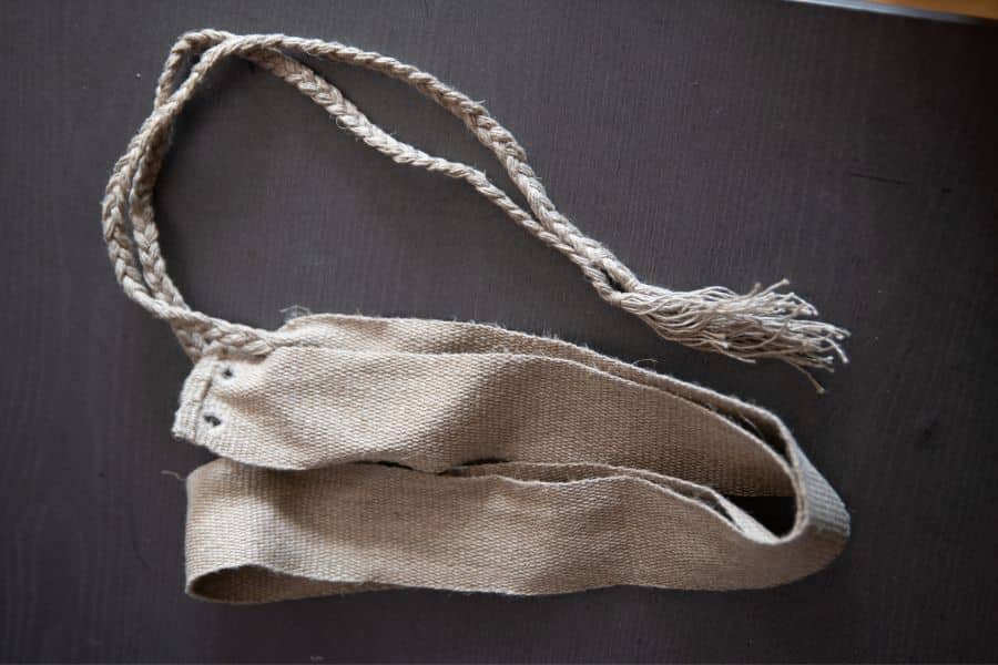 A beige woven fabric strap lies folded on a dark surface beside a long braided cord with a tasselled end. Small stitched tabs and metal eyelets are visible at one end of the strap.