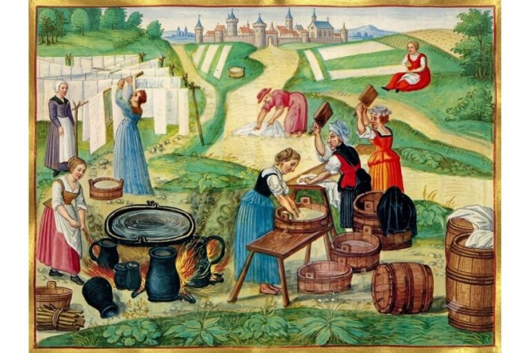 Medieval manuscript scene of peasant women washing and drying laundry outdoors beside a large heated tub wooden barrels and white cloths hanging on a line. A walled town and green fields appear in the background.