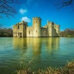 Moated stone castle stands in the middle of a calm lake with bare tree branches framing the scene. The water and strong round towers create a timeless view often associated with English castles.