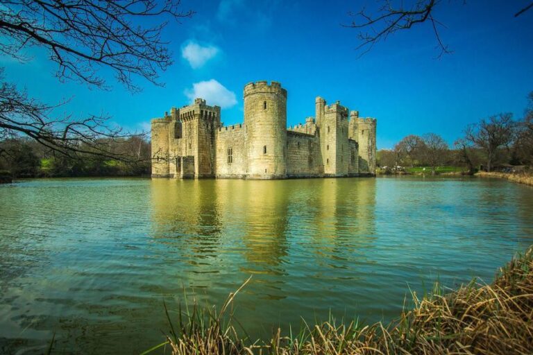 Moated stone castle stands in the middle of a calm lake with bare tree branches framing the scene. The water and strong round towers create a timeless view often associated with English castles.