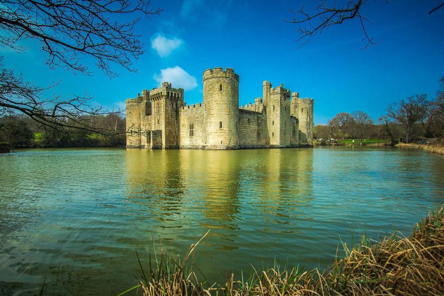 Moated stone castle stands in the middle of a calm lake with bare tree branches framing the scene. The water and strong round towers create a timeless view often associated with English castles.