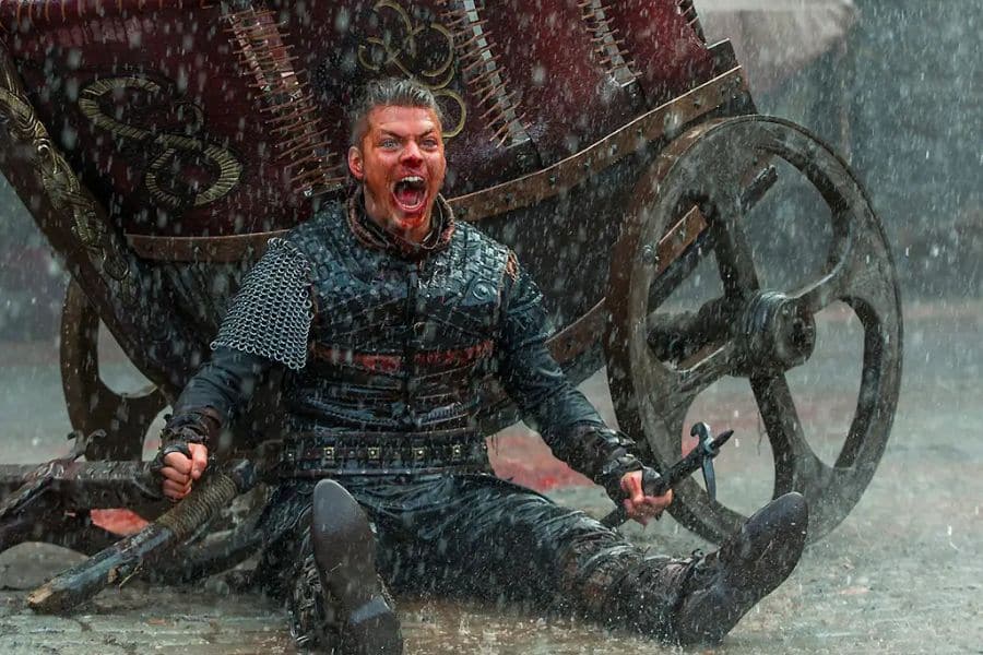 A bloodied Viking warrior in chain mail sits in heavy rain before a damaged wooden wagon while gripping two weapons. His open mouthed yell and soaked armor create a brutal battlefield moment associated with portrayals of Ivar the Boneless.