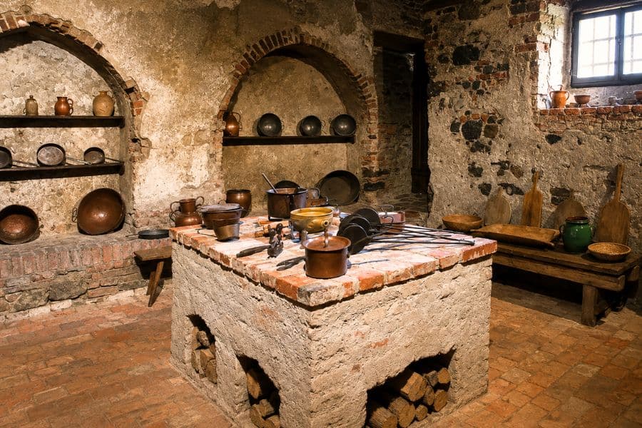 A historic stone kitchen has a large central brick cooking hearth covered with copper pots and long handled utensils. Shelves, benches, and arched wall recesses hold additional cookware used for food preparation.