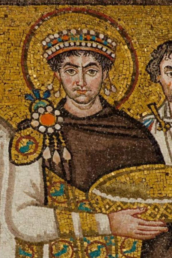 A Byzantine style mosaic depicts a crowned ruler wearing an ornate white robe with gold and jeweled details. A golden halo frames his head while he holds a large ceremonial vessel.