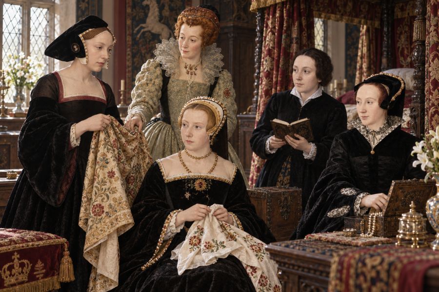 Five women in elaborate Tudor era clothing gather in a richly decorated chamber while sewing reading and handling embroidered fabric. The domestic court setting shows Ladies-in-Waiting together among carved furniture tapestries flowers and patterned textiles.