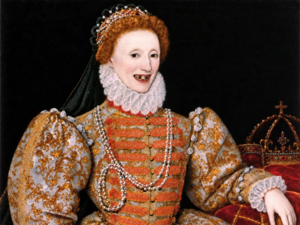Portrait of a smiling red haired royal woman with visibly darkened and missing teeth. She wears an elaborate gold embroidered gown with puffed sleeves and pearl necklaces. A jeweled crown rests beside her against a black background.