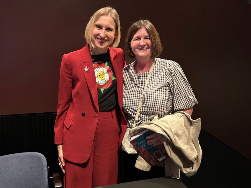 Two smiling women pose together at an indoor event. One wears a red trouser suit over a black shirt featuring a Tudor rose while the other wears a black and white checked top and holds a book and beige jacket.