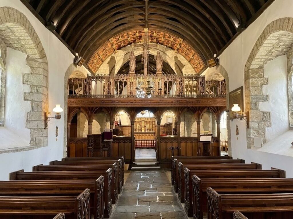 Interior of a historic church with rows of wooden pews leading toward an example of  the ornate rood lofts and altar. The richly carved rood loft spans the nave beneath a dark timber vaulted ceiling.