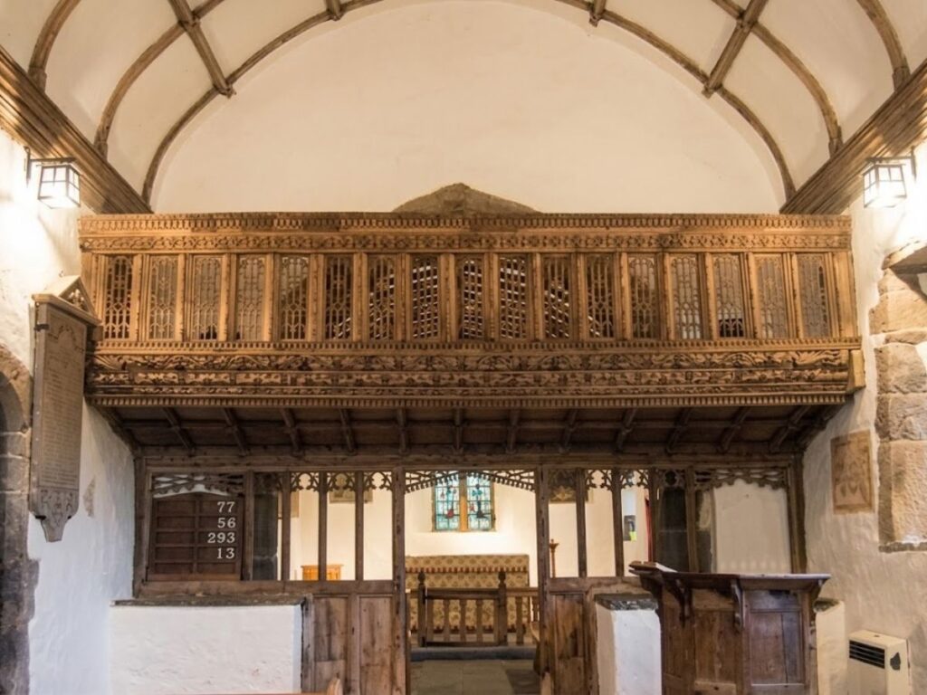 Large carved wooden rood loft spanning the interior of a historic church beneath a ribbed ceiling. Detailed Gothic tracery covers the upper gallery while a wooden screen and altar sit below.