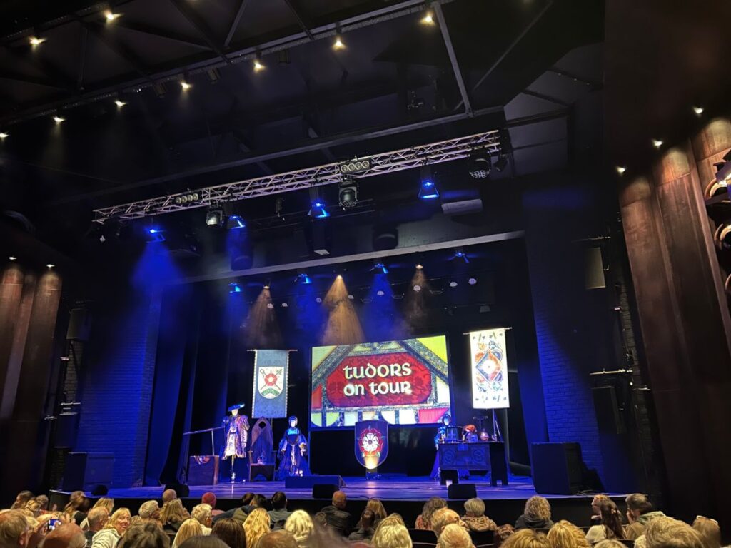 A theatre stage illuminated by blue and gold spotlights features Tudor costumes and banners with a large screen displaying "Tudors on Tour". An audience gathers for a live historical performance exploring Tudor history and Tudor myths.