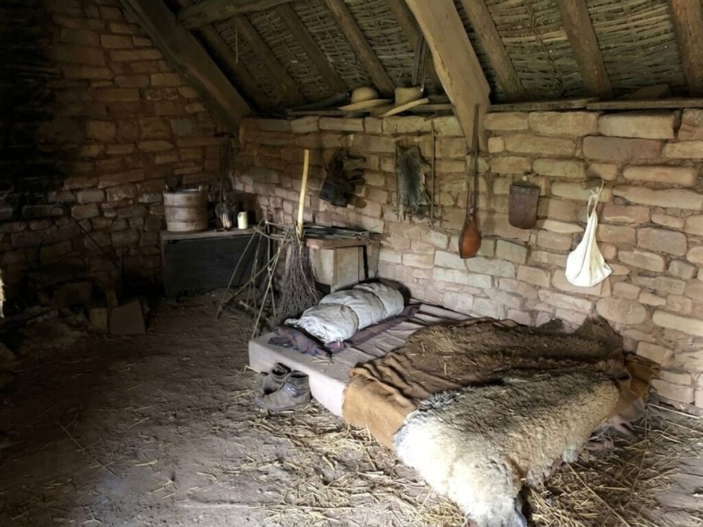 Simple medieval bed made from a low wooden frame with rough blankets and animal skins inside a small stone dwelling. The basic design contrasts with the more elaborate medieval beds used by wealthier households.