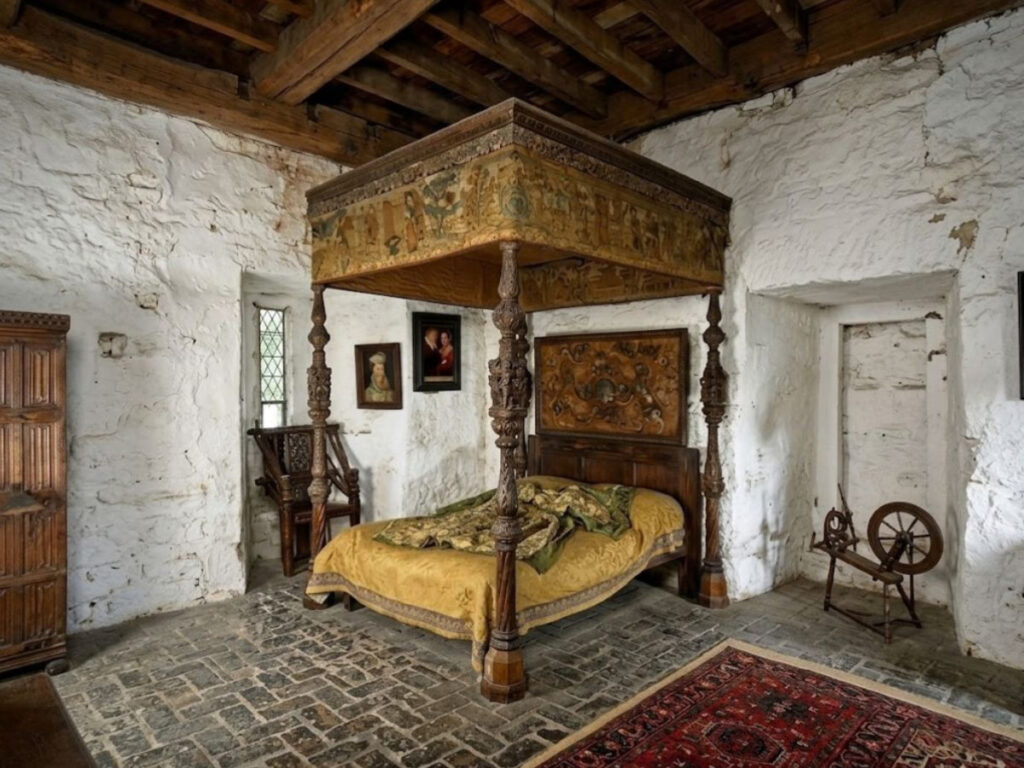 Carved medieval canopy bed covered with yellow and green bedding in a whitewashed stone bedroom. This decorative example of medieval beds is surrounded by a wooden chair spinning wheel portraits and a patterned rug.