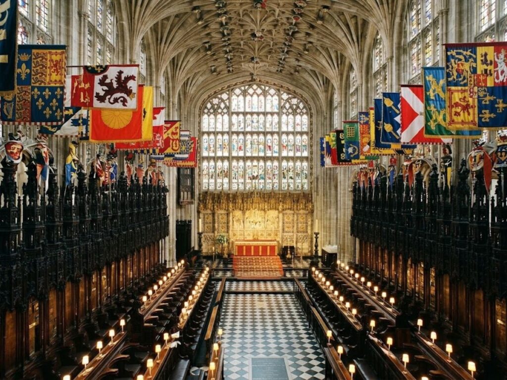 Interior of St Georges Chapel at Windsor Castle where Henry VIII is buried. Colorful heraldic banners hang above dark wooden choir stalls beneath an intricately carved vaulted ceiling with a large stained glass window overlooking the altar.
