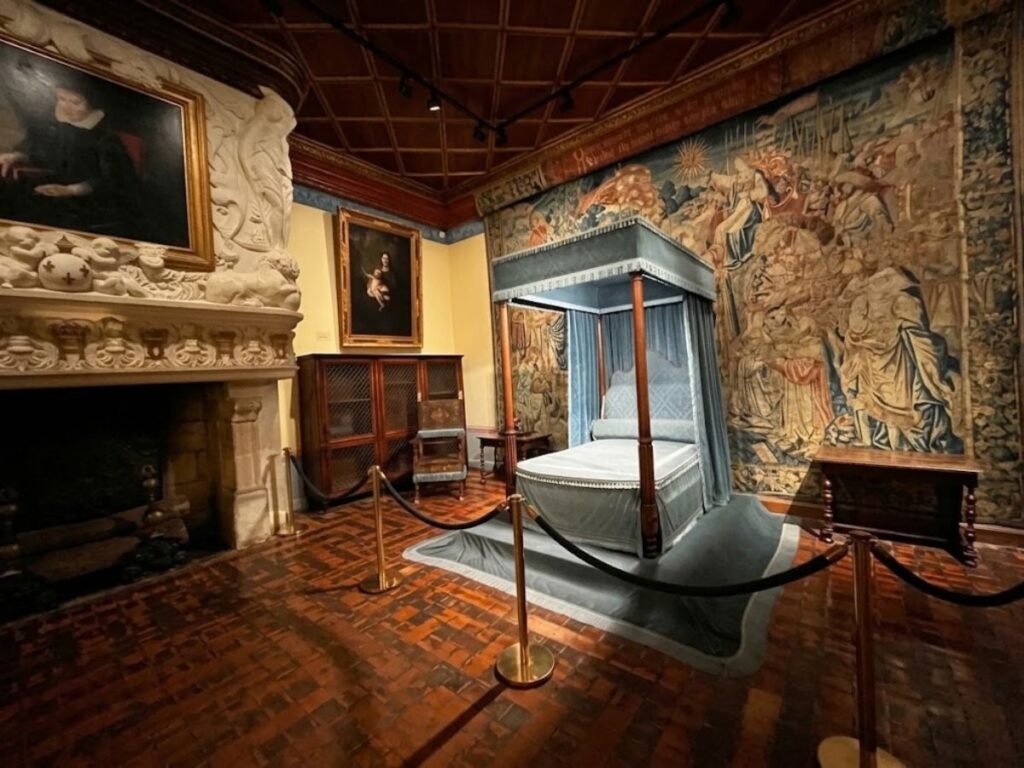 Formal medieval bed with a tall blue canopy displayed in a richly decorated chamber surrounded by large narrative tapestries. The elaborate setting shows the luxury possible in medieval beds made for wealthy households.
